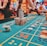 A casino scene with a roulette table features prominently, covered in green felt with colorful numbers and betting squares. Several stacks of poker chips are placed on different numbers, and the hands of several people are visible as they interact with the table. In the background, there are slot machines and more people, creating a vibrant and busy atmosphere.